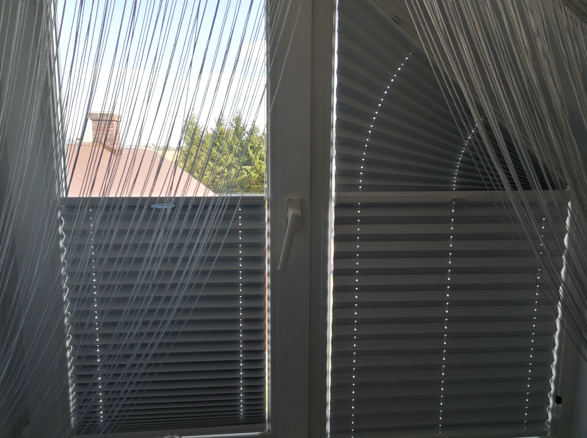 Okno z zamontowanymi plisami w kolorze szarym, z widokiem na dach domu i drzewa, zasłonięte dodatkowo dekoracyjną zasłoną sznurkową.