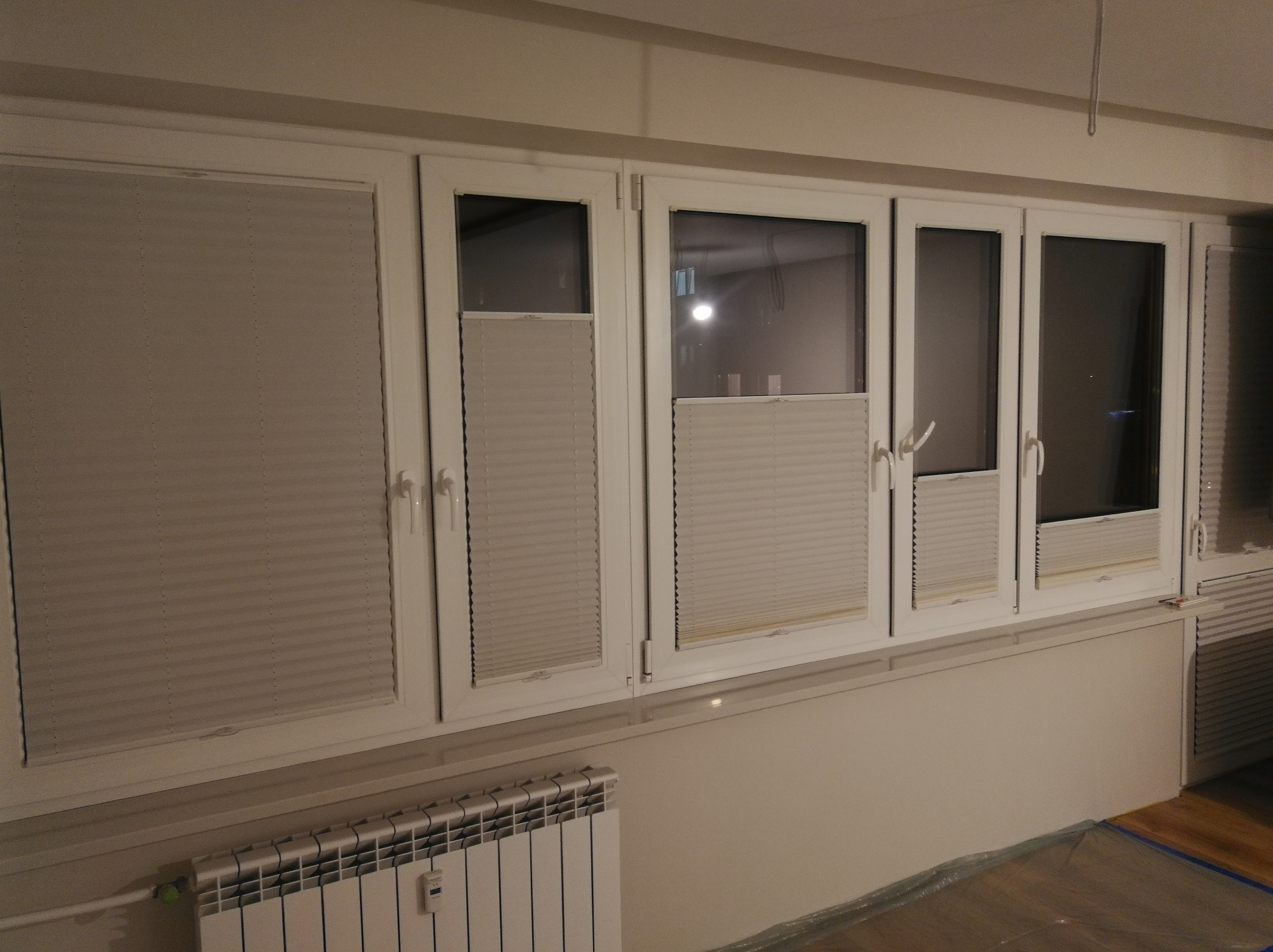 Szereg białych okien z zamontowanymi jasnoszarymi plisami, niektóre z nich częściowo zasłonięte, widoczny grzejnik pod oknem i folia ochronna na podłodze.