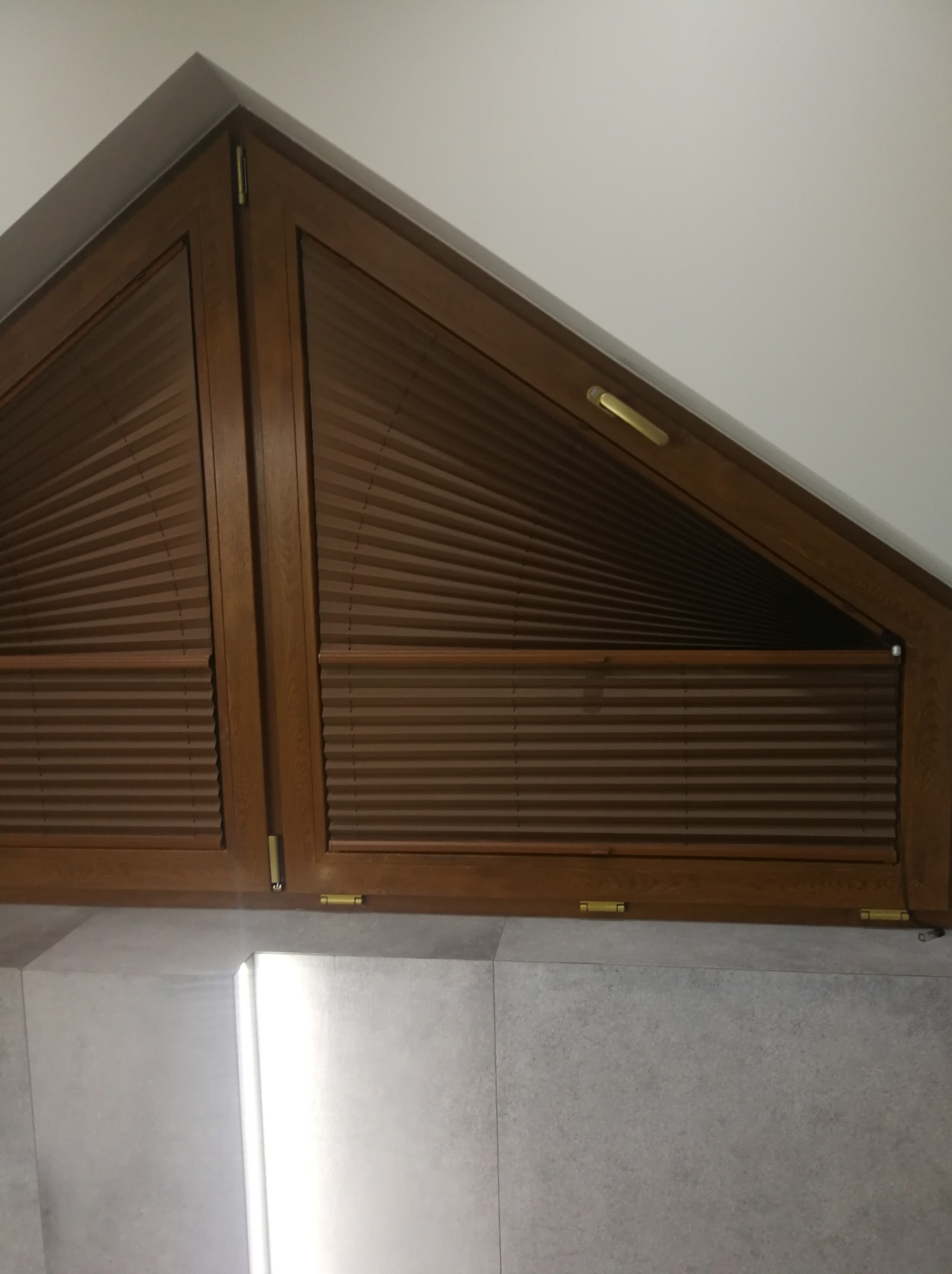 Brązowe plisy zamontowane w oknie o nietypowym, trójkątnym kształcie, drewniana rama okienna, widok z dołu.