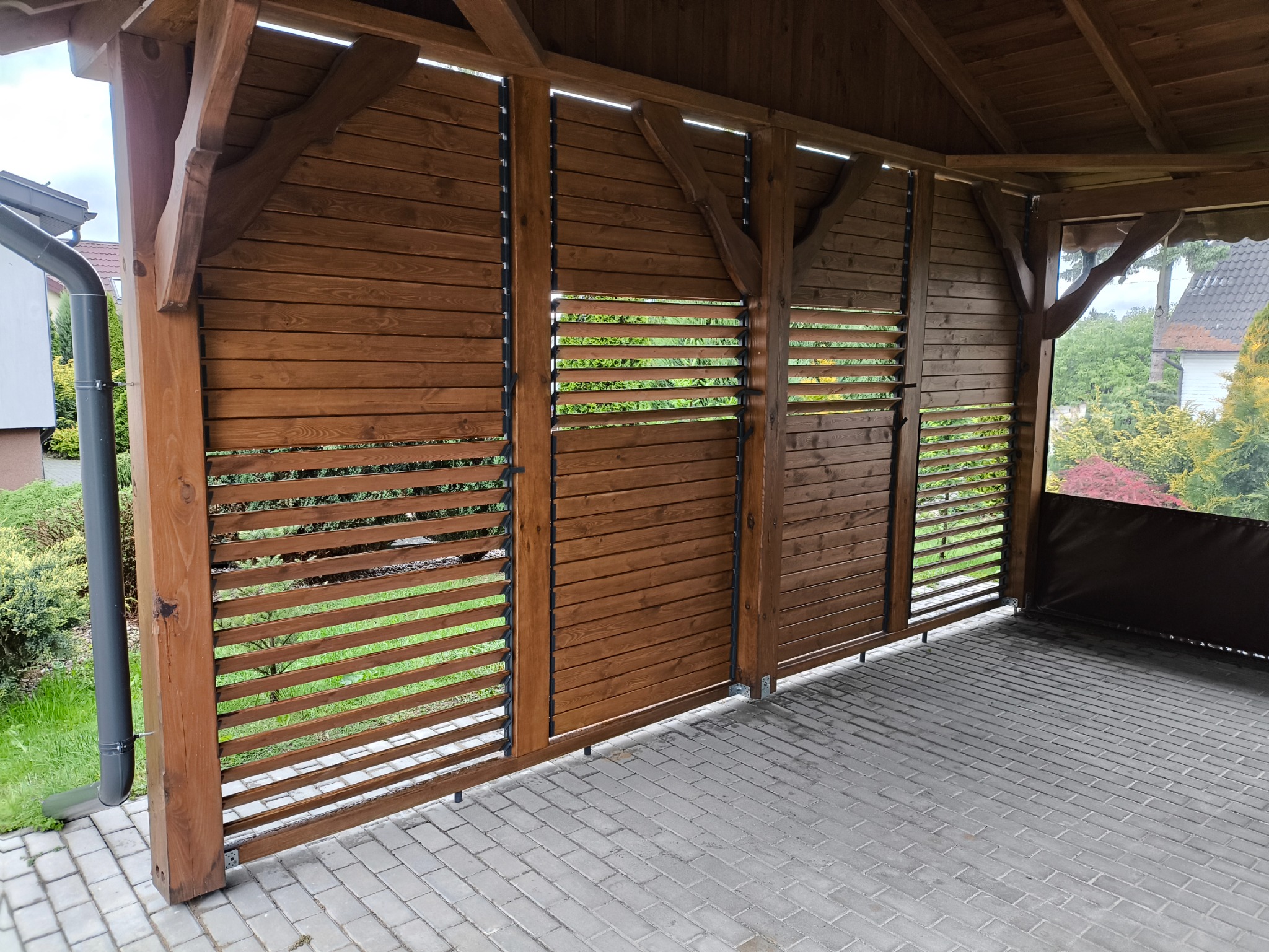 Drewniana pergola z ażurowymi panelami, widok z wnętrza. Brązowe drewno, szara kostka brukowa, zieleń w tle. Elementy konstrukcyjne z widocznymi łączeniami.