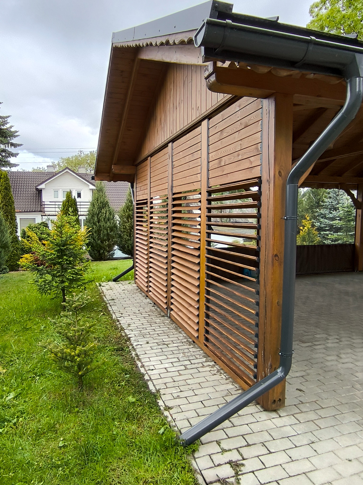 Drewniana pergola z lamelowymi panelami bocznymi i systemem odprowadzania wody. Widok z zewnątrz, zieleń i kostka brukowa w tle.