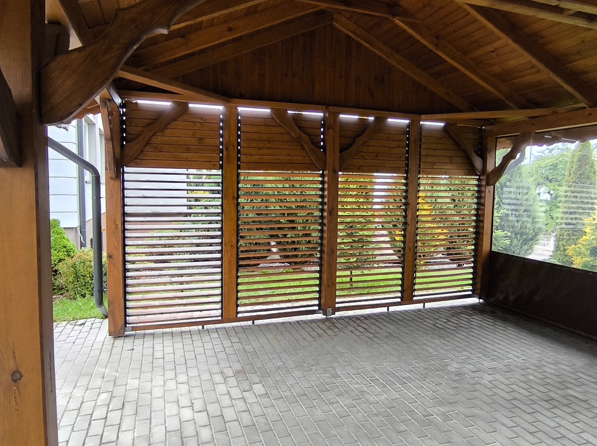 Drewniana pergola z lamelowymi panelami bocznymi, zapewniającymi prywatność i cień. Widoczna kostka brukowa i zieleń w tle. Konstrukcja z belek w ciepłym odcieniu brązu.