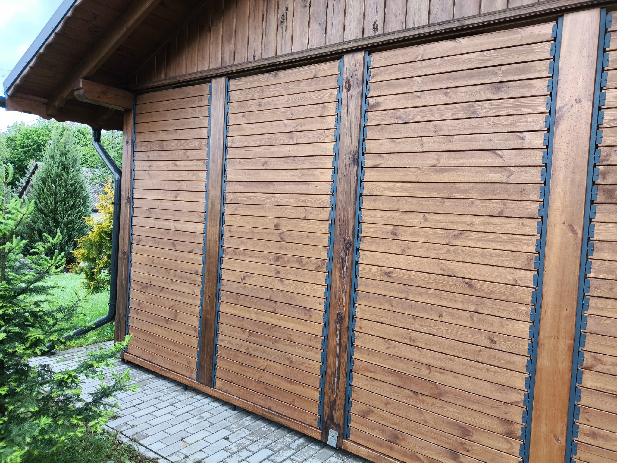 Drewniana zabudowa balkonu z przesuwnymi panelami, widok z zewnątrz, zieleń w tle. Naturalne oświetlenie podkreśla strukturę drewna.