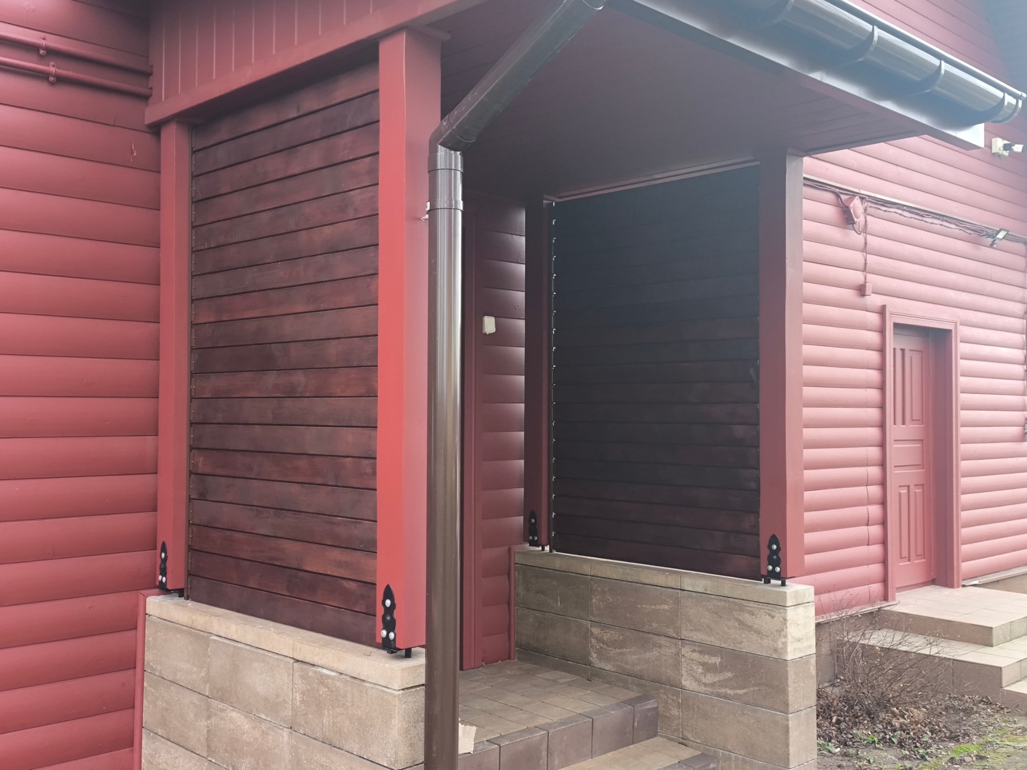 Zabudowa tarasu drewnianymi lamelami w kolorze ciemnego brązu, z widocznymi elementami mocującymi. Budynek w odcieniach czerwieni. Widoczne schody.