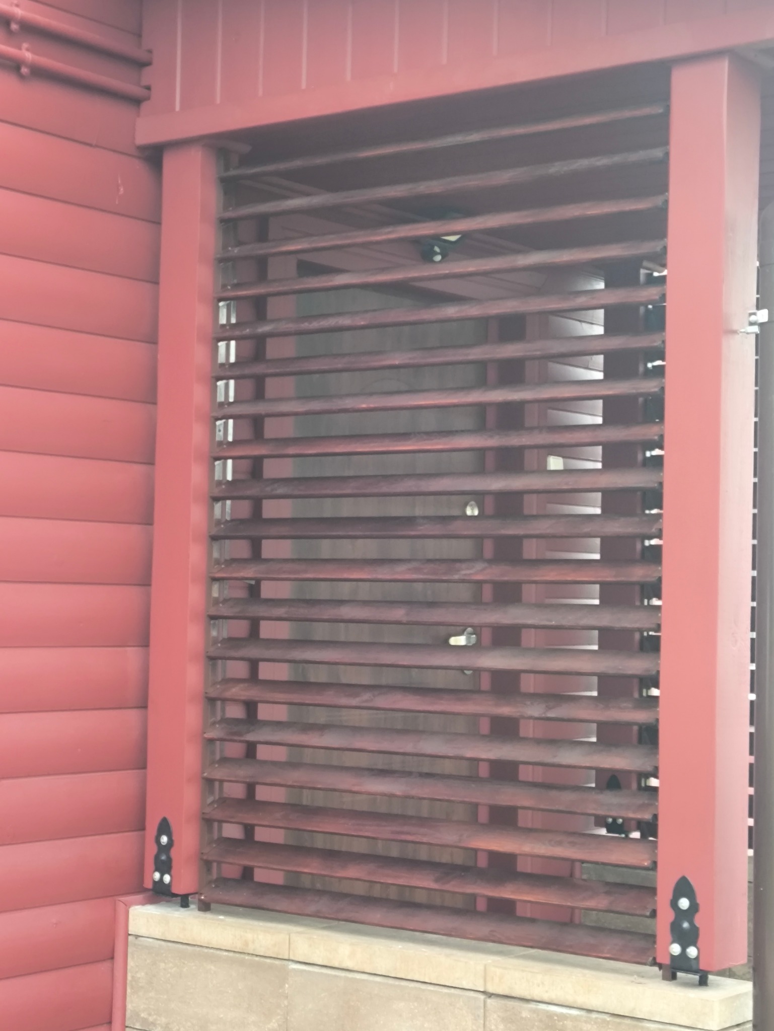 Czerwona, pozioma przesłona okienna z drewna, zamontowana na tle ściany z desek w tym samym kolorze. Widoczne detale mocowania i konstrukcji.