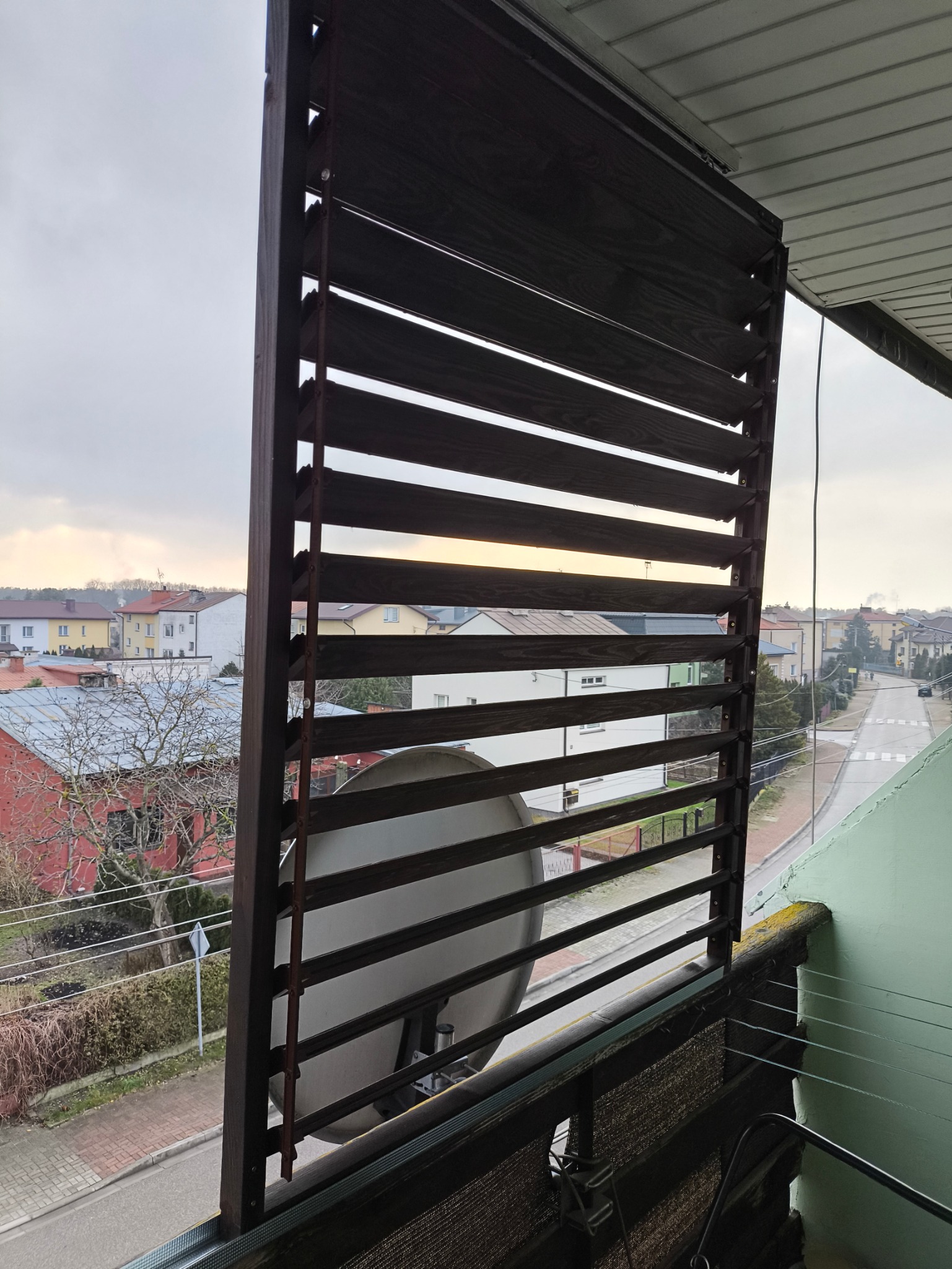 Drewniane żaluzje balkonowe w kolorze ciemnego brązu, zamontowane na balkonie z widokiem na ulicę i zabudowę miejską w pochmurny dzień.