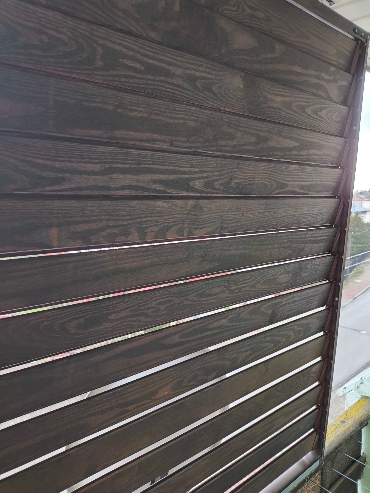 Ciemne, drewniane żaluzje zewnętrzne zamontowane na balkonie, widoczne z bliska pod kątem. Widoczne słoje drewna i uliczny krajobraz w tle.