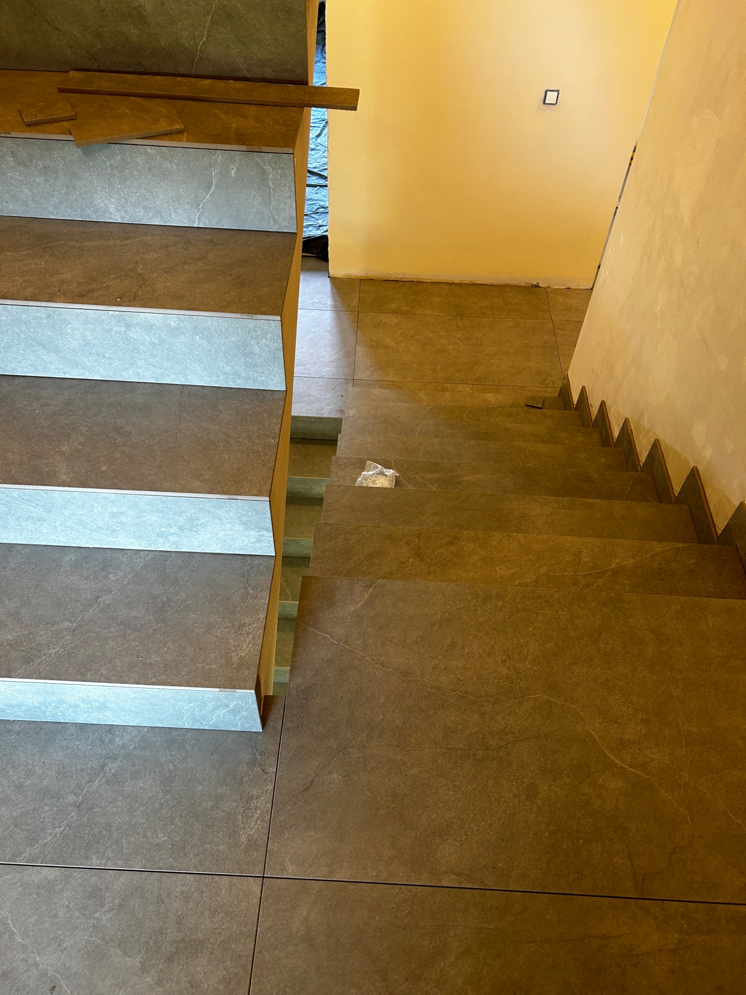 Schody wyłożone szarymi płytkami imitującymi kamień, z widocznymi aluminiowymi listwami krawędziowymi, częściowo zabezpieczone folią malarską.