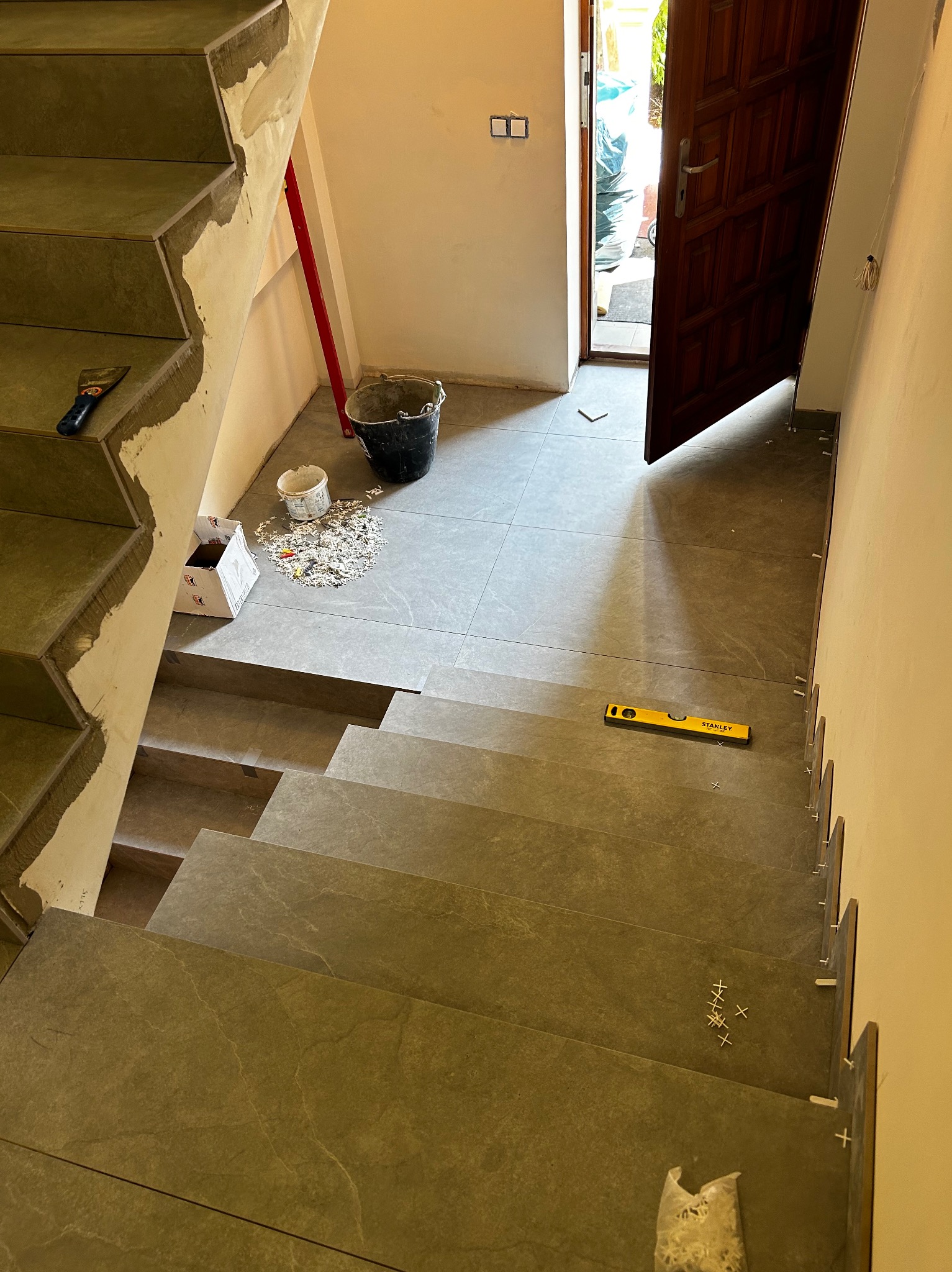 Schody w trakcie wykańczania szarą płytką imitującą kamień, widoczne narzędzia glazurnicze, poziomica Stanley, krzyżyki dystansowe i pozostałości zaprawy.