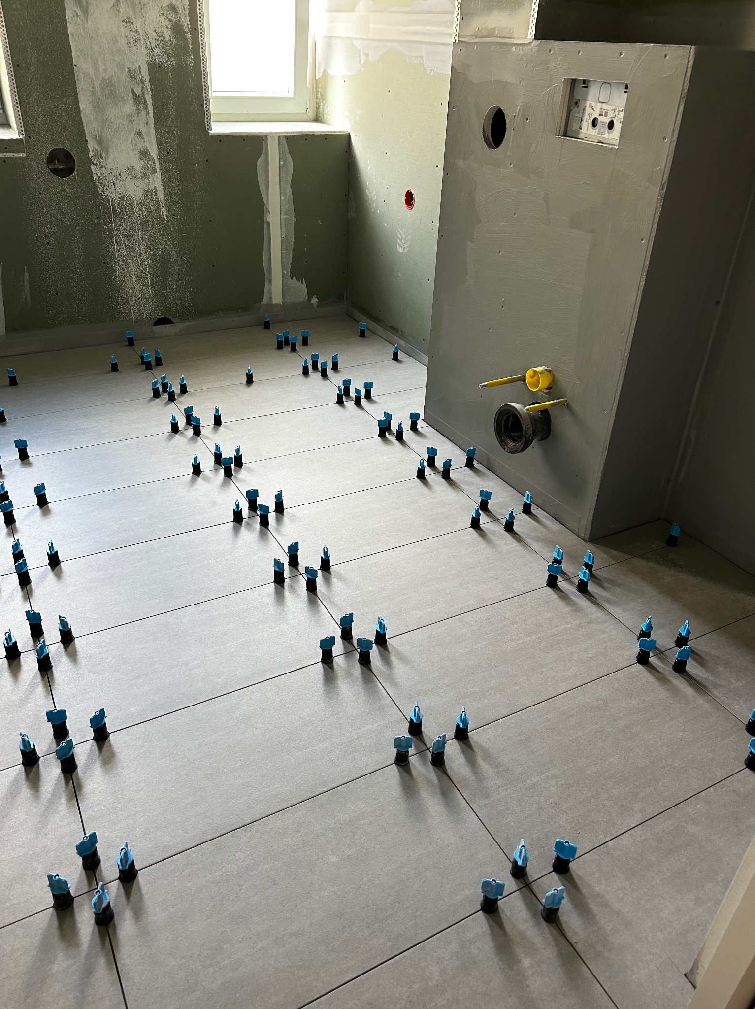 Szare płytki podłogowe w łazience, wyrównane za pomocą niebieskich klipsów poziomujących, widoczne przygotowane ściany z płyt gipsowych i instalacja podtynkowa WC z wystającymi żółtymi rurami.