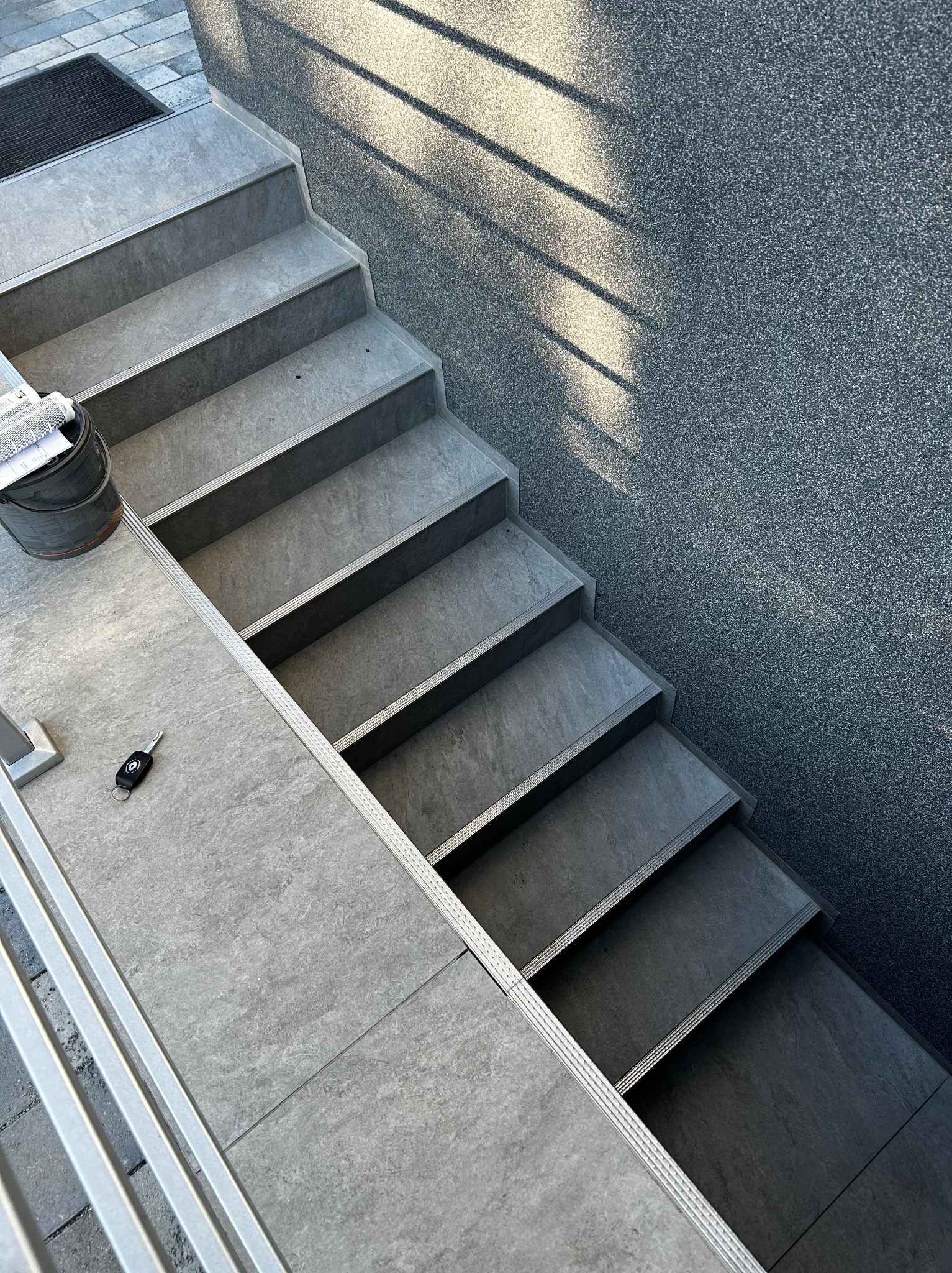 Szare schody wyłożone płytkami, widok z góry, z boku ściana o chropowatej strukturze, na pierwszym planie klucze samochodowe i puszka z farbą.