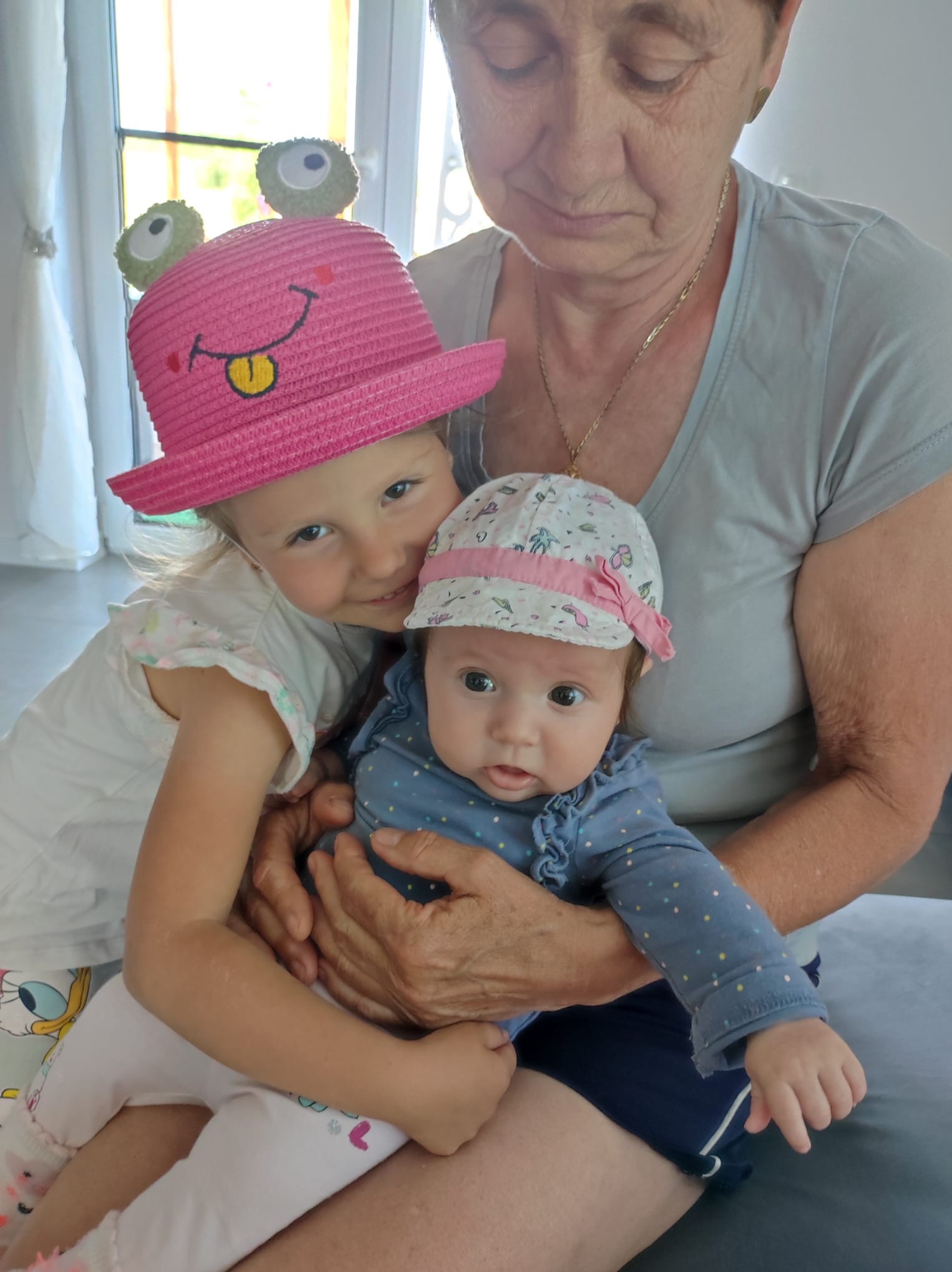 Babcia trzyma wnuczkę w kapeluszu, a druga wnuczka w różowym kapeluszu z żabą przytula się do niej.