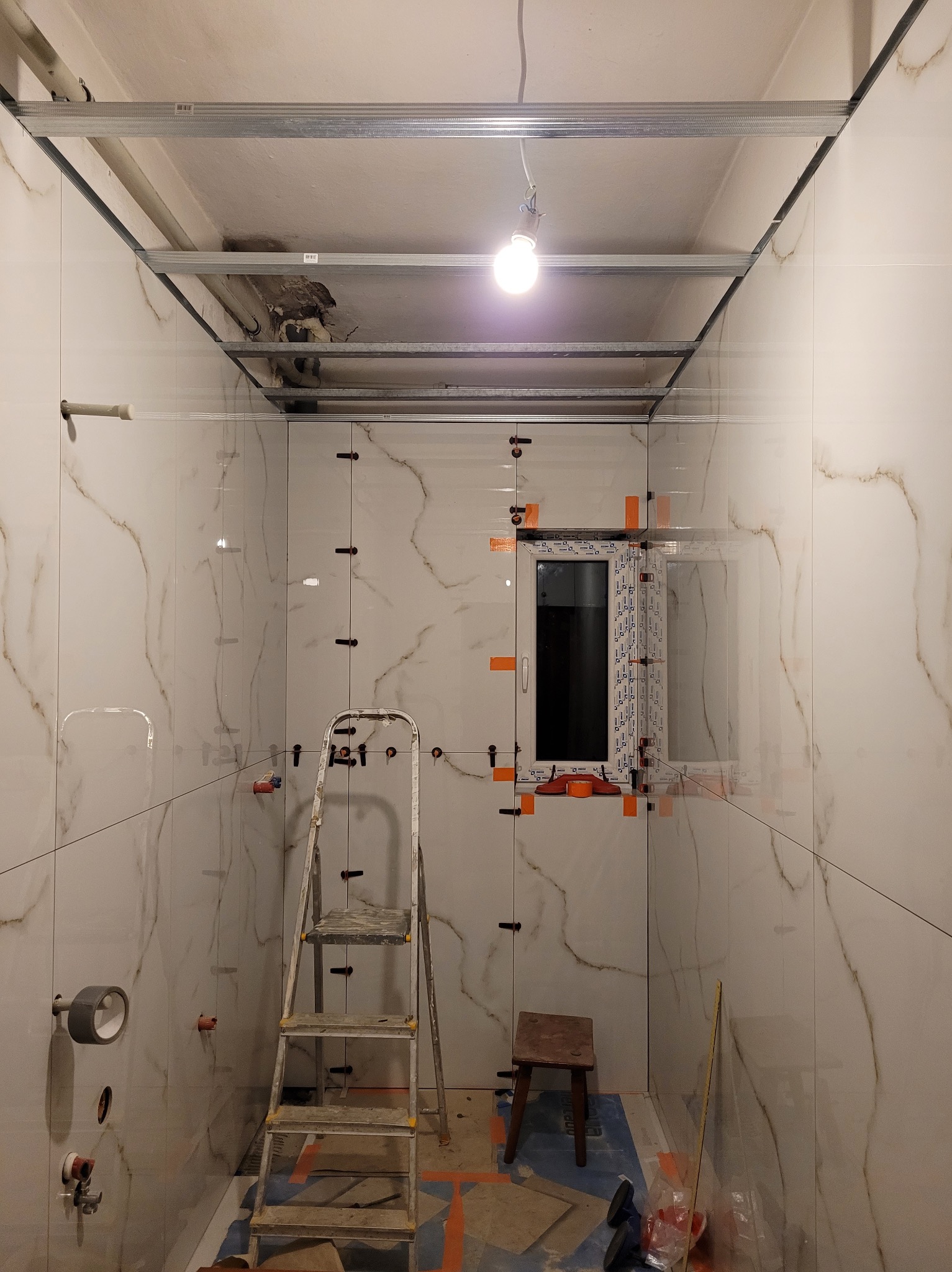 Łazienka w trakcie remontu: ułożone marmurowe płytki z krzyżykami dystansowymi, widoczna drabina, drewniane krzesło,  metalowa konstrukcja sufitu podwieszanego, zamontowane okno.