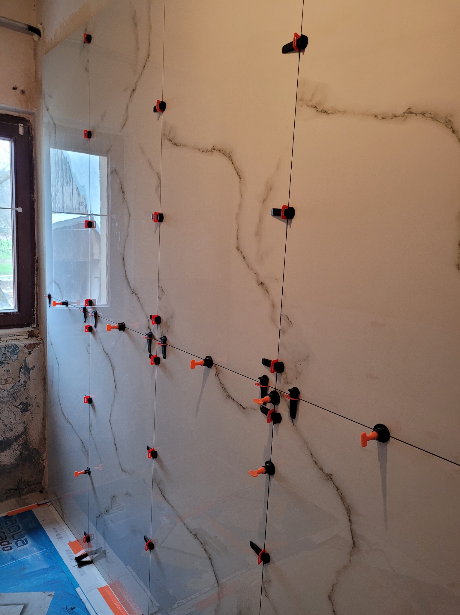 Ściana z marmurkowych płytek w trakcie montażu, z użyciem systemu poziomowania płytek z pomarańczowymi klinami i czarnymi klipsami, widoczne okno i fragment podłogi z folią ochronną.