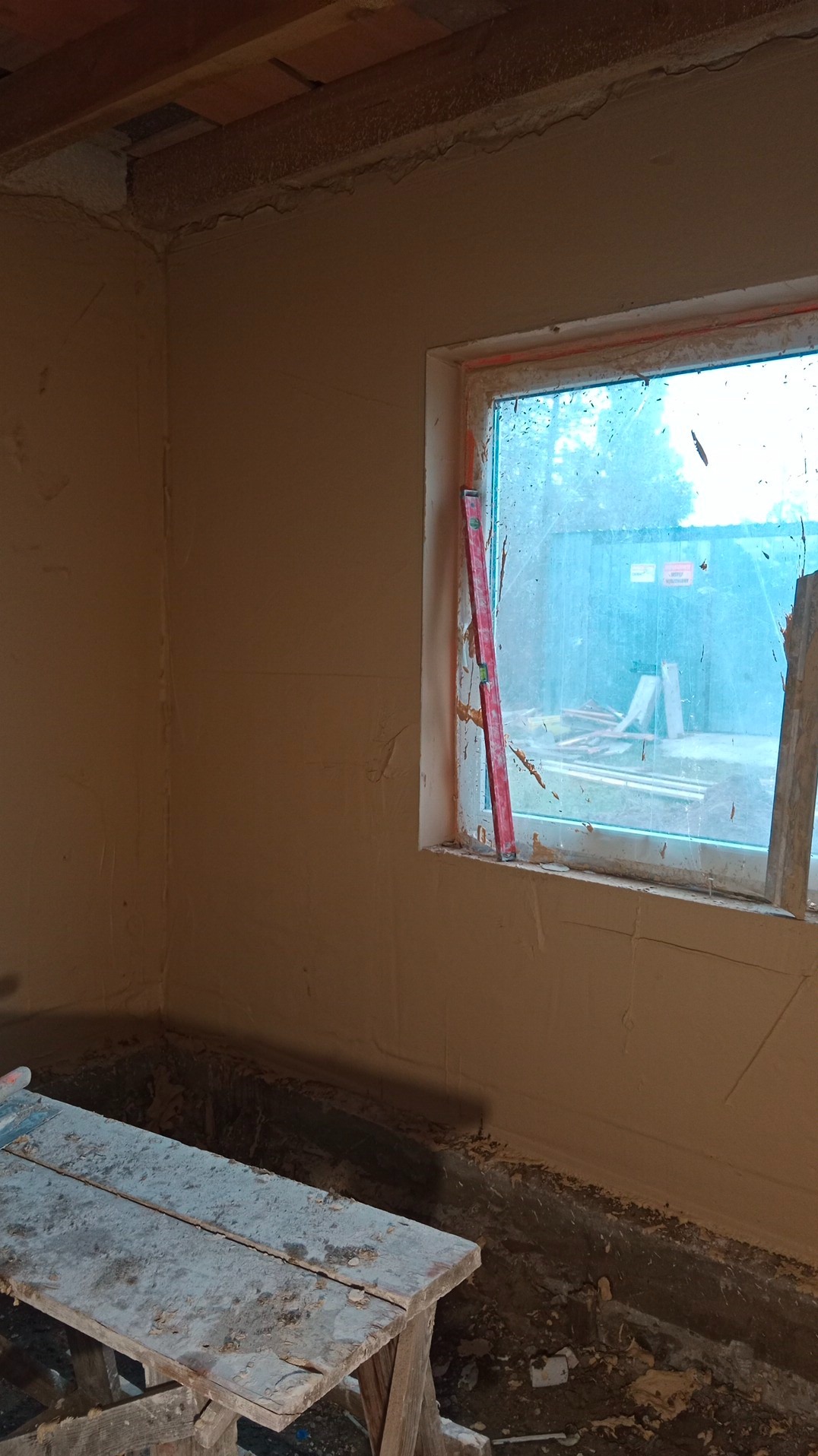 Wnętrze pokoju w trakcie prac wykończeniowych, ściany pokryte świeżym tynkiem, przy oknie pionowy poziomica, widoczny fragment drewnianego stołu roboczego i nieporządek na podłodze.