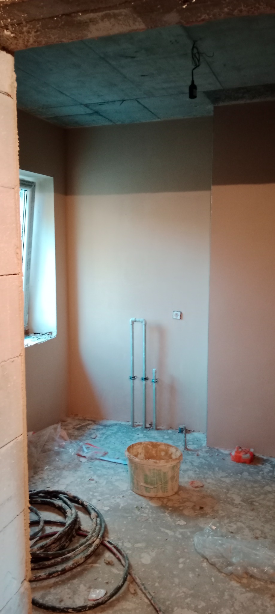 Wnętrze w trakcie prac wykończeniowych: ściany częściowo pokryte świeżym jasnym tynkiem, widoczne instalacje wodne, wiadro z resztkami zaprawy i zwinięte kable na podłodze zabezpieczonej folią.