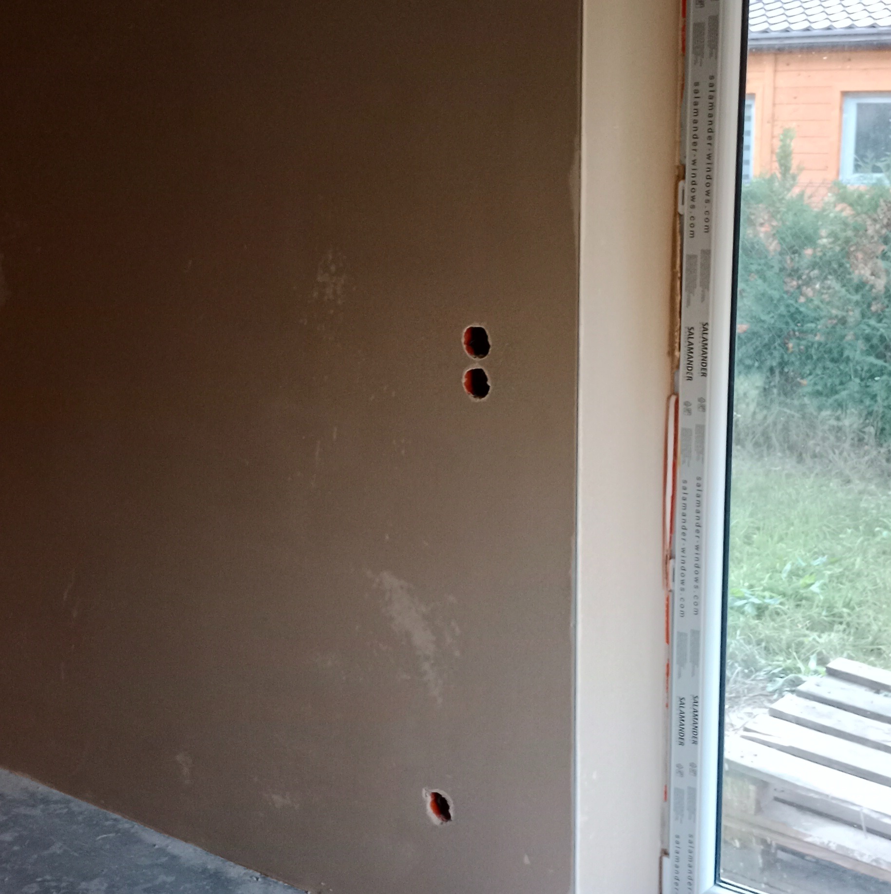 Świeżo nałożony, gładki tynk na ścianie, z widocznymi otworami na gniazdka elektryczne i ramą okienną z logo producenta Salamander.