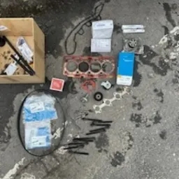 Rozłożone części samochodowe na betonowej posadzce, widoczne uszczelki, głowica silnika, pudełka z częściami zamiennymi oraz narzędzia w drewnianej skrzynce, u dołu kadru stopy w trampkach.