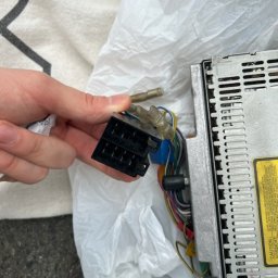 Mobilny Mechanik Elektryk Samochodowy Gdańsk Gdynia Sopot - Czarny konektor w dłoni osoby, obok radia samochodowego z widocznymi przewodami, na tle plastikowej torby i fragmentu materiału.