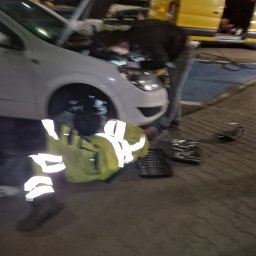 Mobilny Mechanik Elektryk Samochodowy Gdańsk Gdynia Sopot - Mechanik w odblaskowej kamizelce naprawia biały samochód z otwartą maską, obok leżą narzędzia, w tle żółty bus.