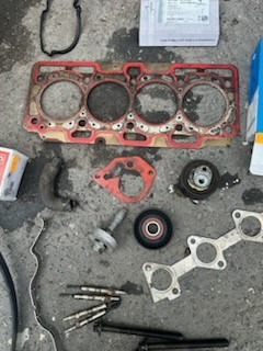 Zestaw używanych części samochodowych na betonowej posadzce: uszczelka głowicy silnika z resztkami oleju, świece żarowe, fragmenty przewodów i inne drobne elementy mechaniczne, pudełka po częściach...