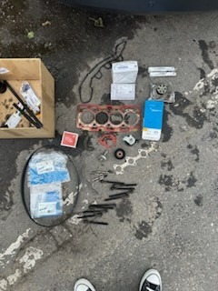 Rozłożone części samochodowe na betonowej posadzce, widoczne uszczelki, głowica silnika, pudełka z częściami zamiennymi oraz narzędzia w drewnianej skrzynce, u dołu kadru stopy w trampkach.