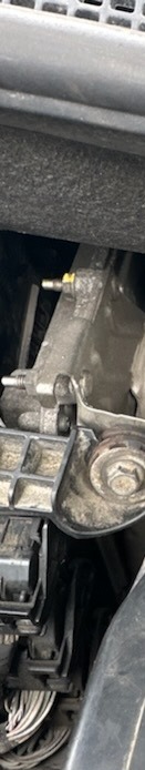 Szczegółowy widok na brudny, metalowy element mechaniczny w komorze silnika samochodowego, z widocznymi przewodami i śrubami.