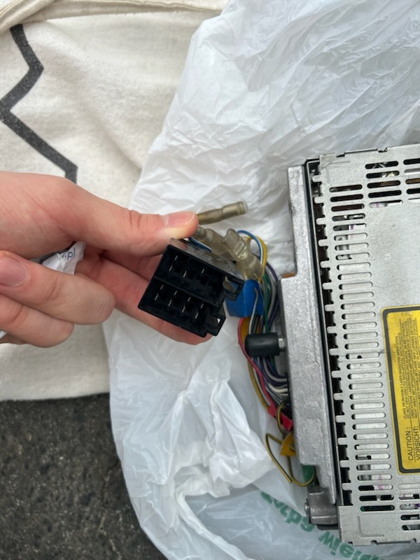 Czarny konektor w dłoni osoby, obok radia samochodowego z widocznymi przewodami, na tle plastikowej torby i fragmentu materiału.
