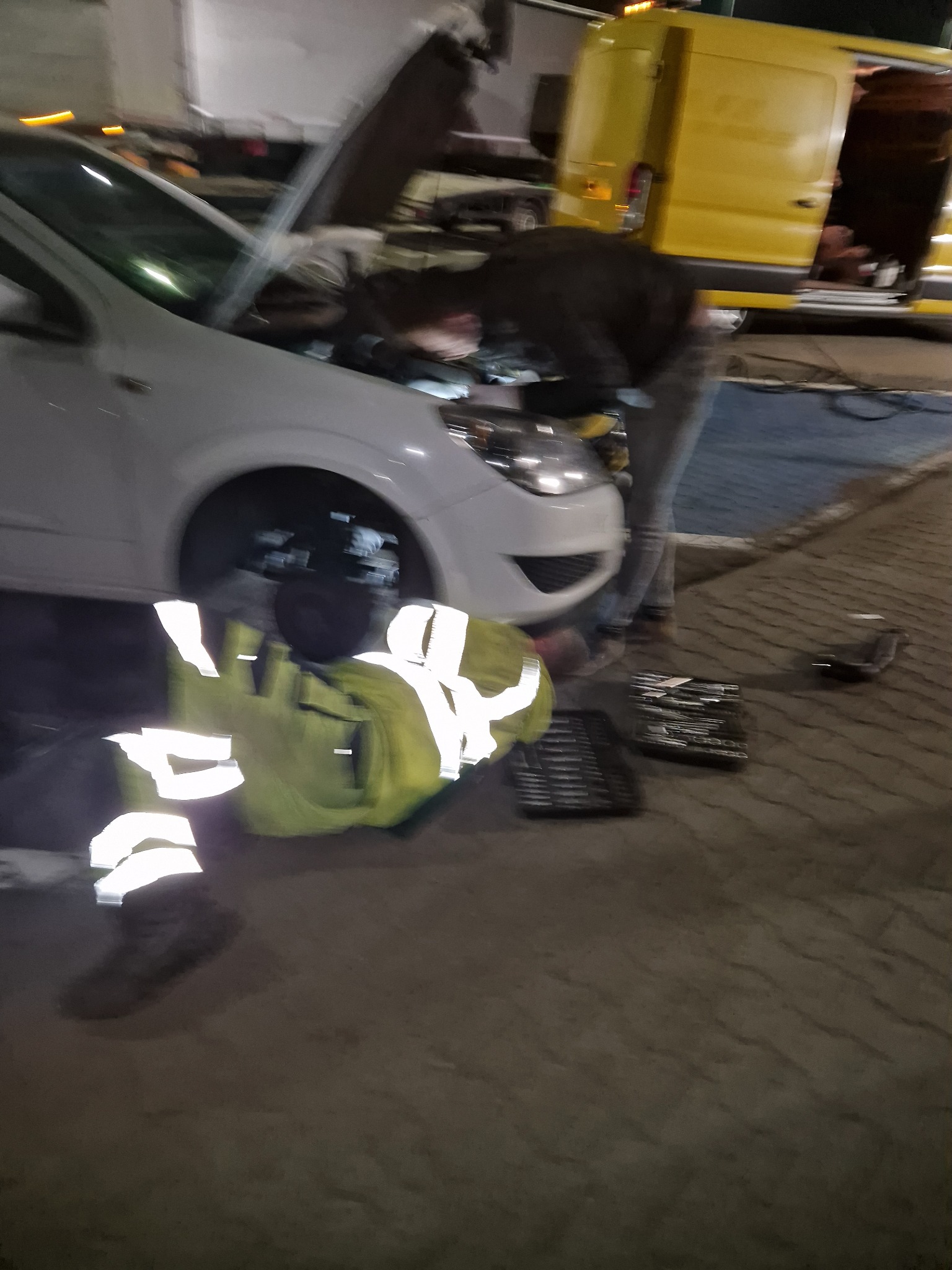 Mechanik w odblaskowej kamizelce naprawia biały samochód z otwartą maską, obok leżą narzędzia, w tle żółty bus.