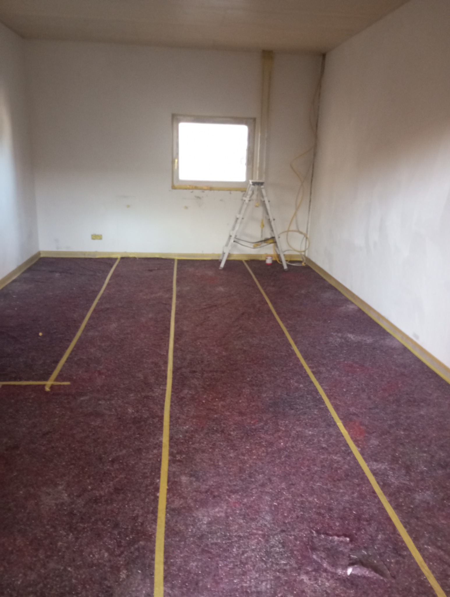 Pokój w trakcie przygotowań do prac wykończeniowych, zabezpieczona podłoga z wyznaczonymi liniami, drabina oparta o ścianę przy oknie.