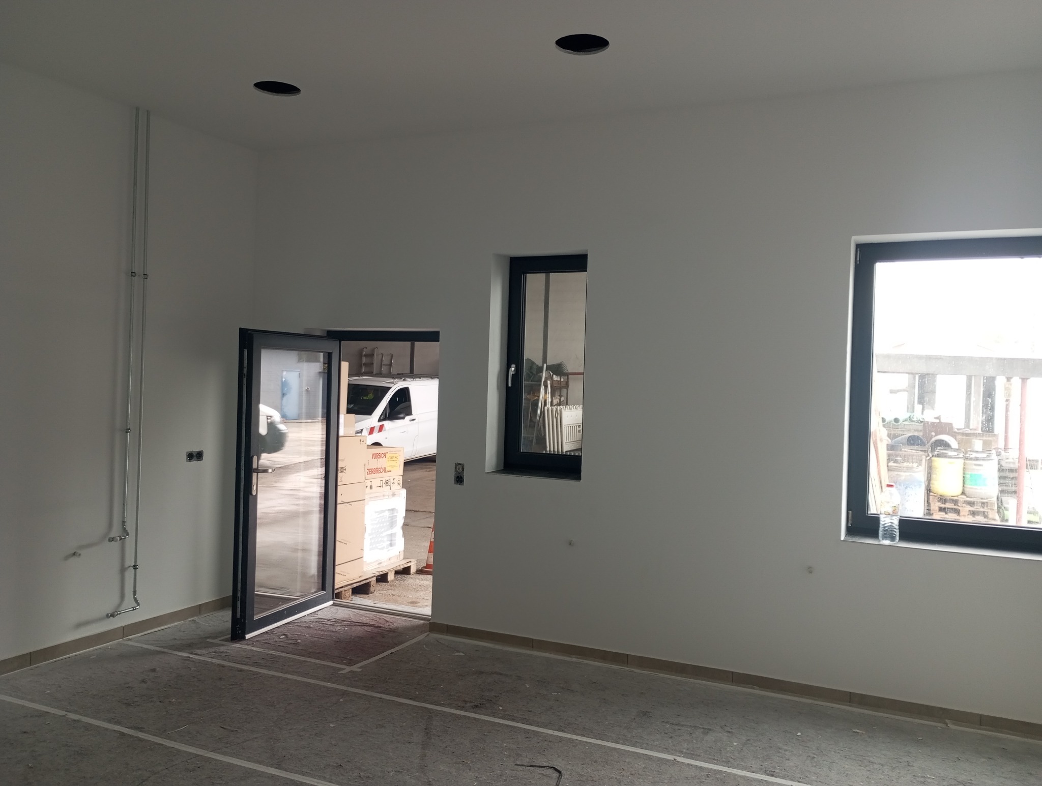 Wnętrze świeżo wykończonego pomieszczenia z białymi ścianami, czarnymi ramami okiennymi i otwartymi drzwiami, przez które widać dostawczy samochód i materiały budowlane.