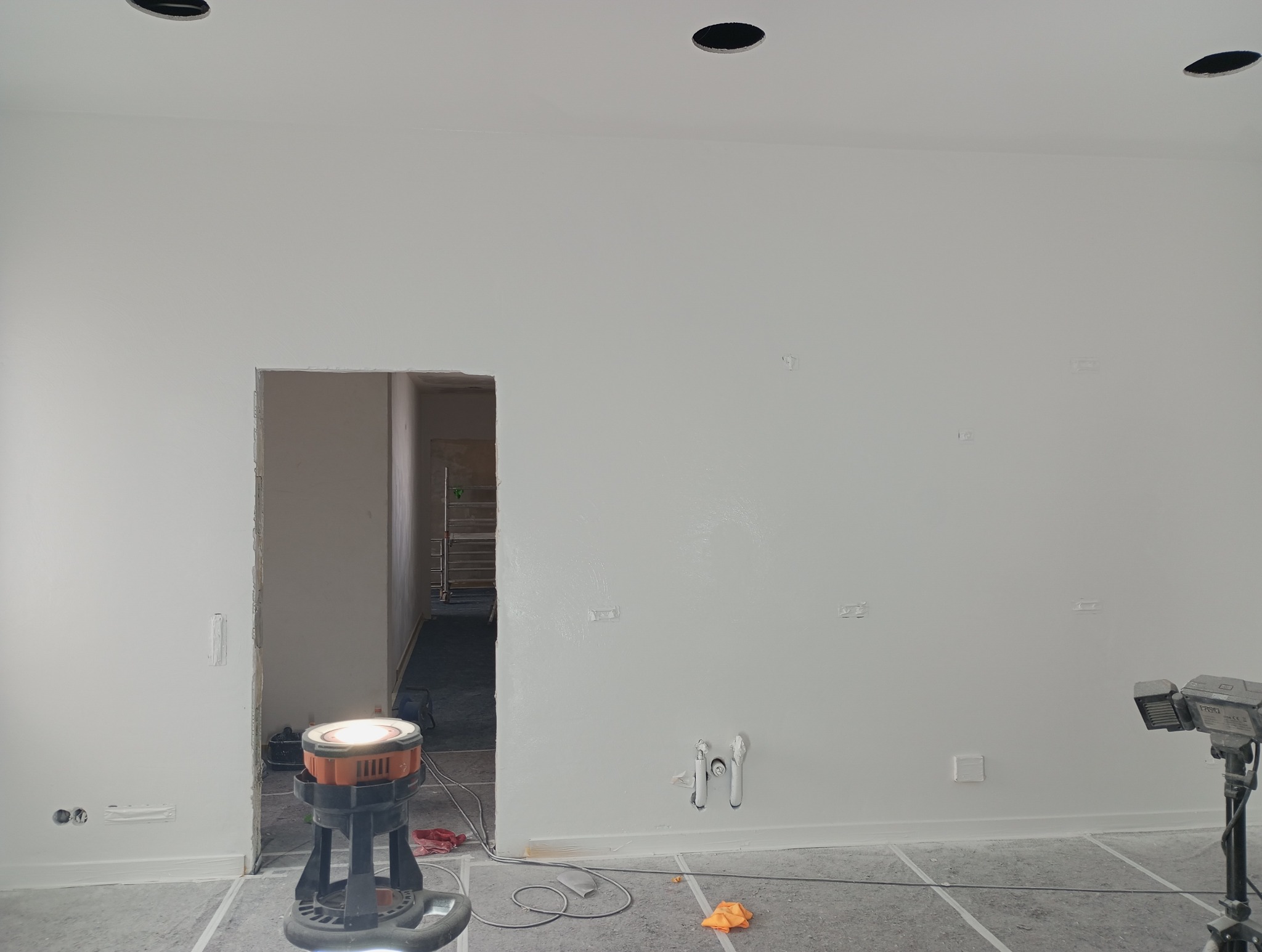 Wnętrze w trakcie remontu: świeżo pomalowana na biało ściana z widocznymi śladami szpachlowania, wystające rury, otwór drzwiowy bez ościeżnicy, halogen budowlany i taśmy malarskie na podłodze.