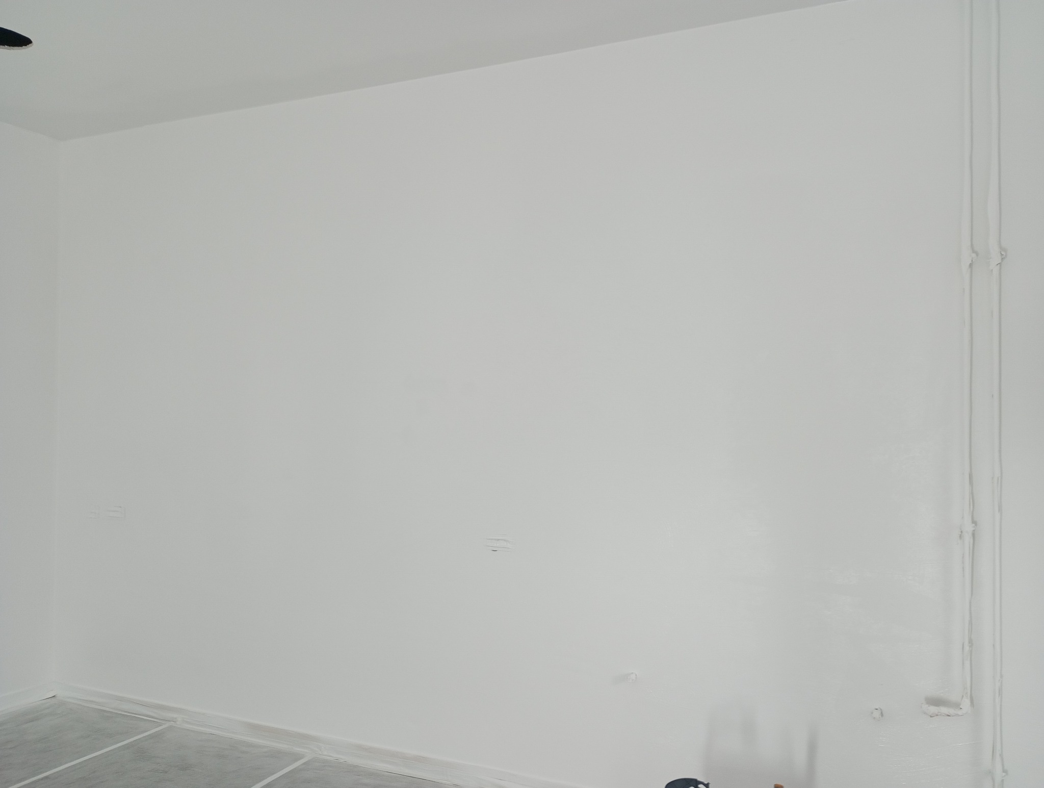 Świeżo pomalowana na biało ściana w pokoju, z widocznymi śladami po szpachlowaniu i zabezpieczoną folią podłogą, ukazująca etap wykończeniowy prac malarskich.