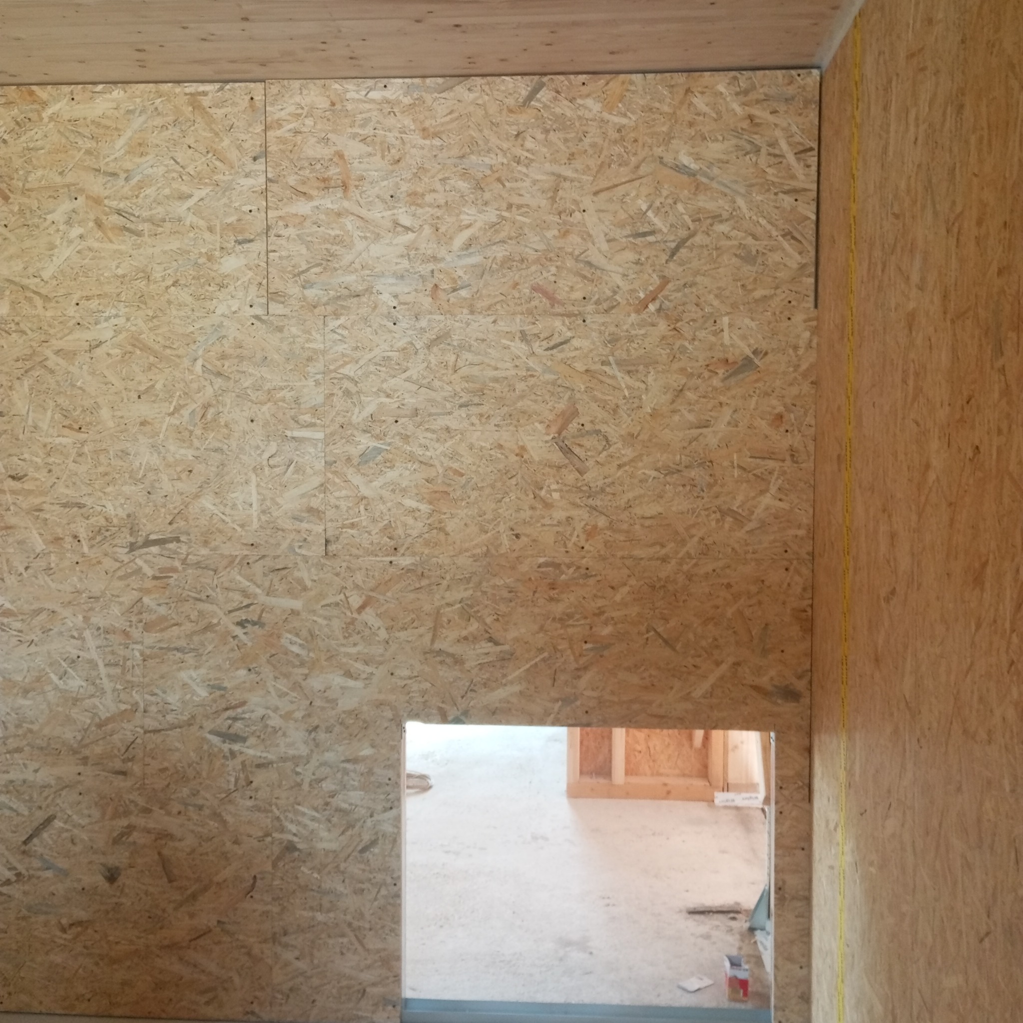 Ściana wewnętrzna wykończona płytą OSB, widoczne łączenia płyt i fragment drewnianego sufitu. W tle otwór drzwiowy ujawnia surowy stan podłogi i konstrukcji.