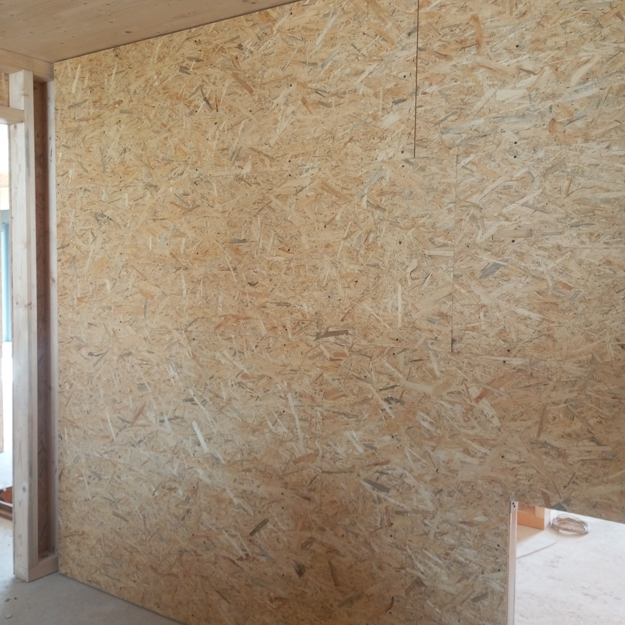 Surowa płyta OSB zamontowana na ścianie z widoczną drewnianą konstrukcją ramy, tworząca przegrodę wewnętrzną w trakcie budowy lub remontu.