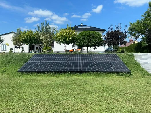 Instalacja paneli słonecznych na trawniku przed domem jednorodzinnym w słoneczny dzień. Widoczne panele ustawione rzędami, w tle dom i drzewa.