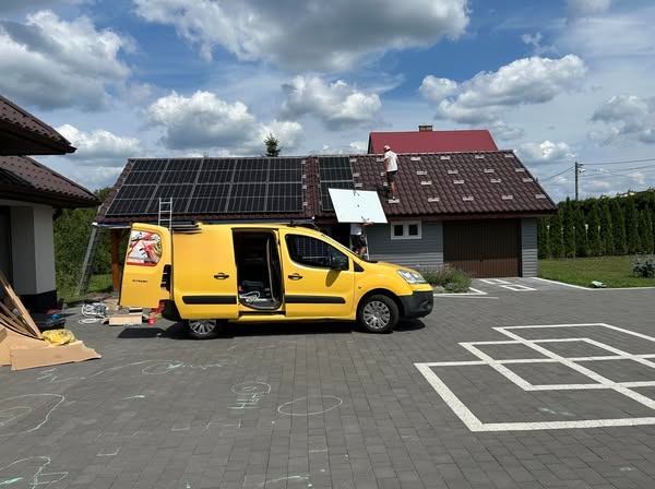 Montaż paneli słonecznych na dachu domu przez dwóch monterów. Żółty samochód dostawczy z otwartymi drzwiami obok. Część paneli już zamontowana. Słoneczny dzień.