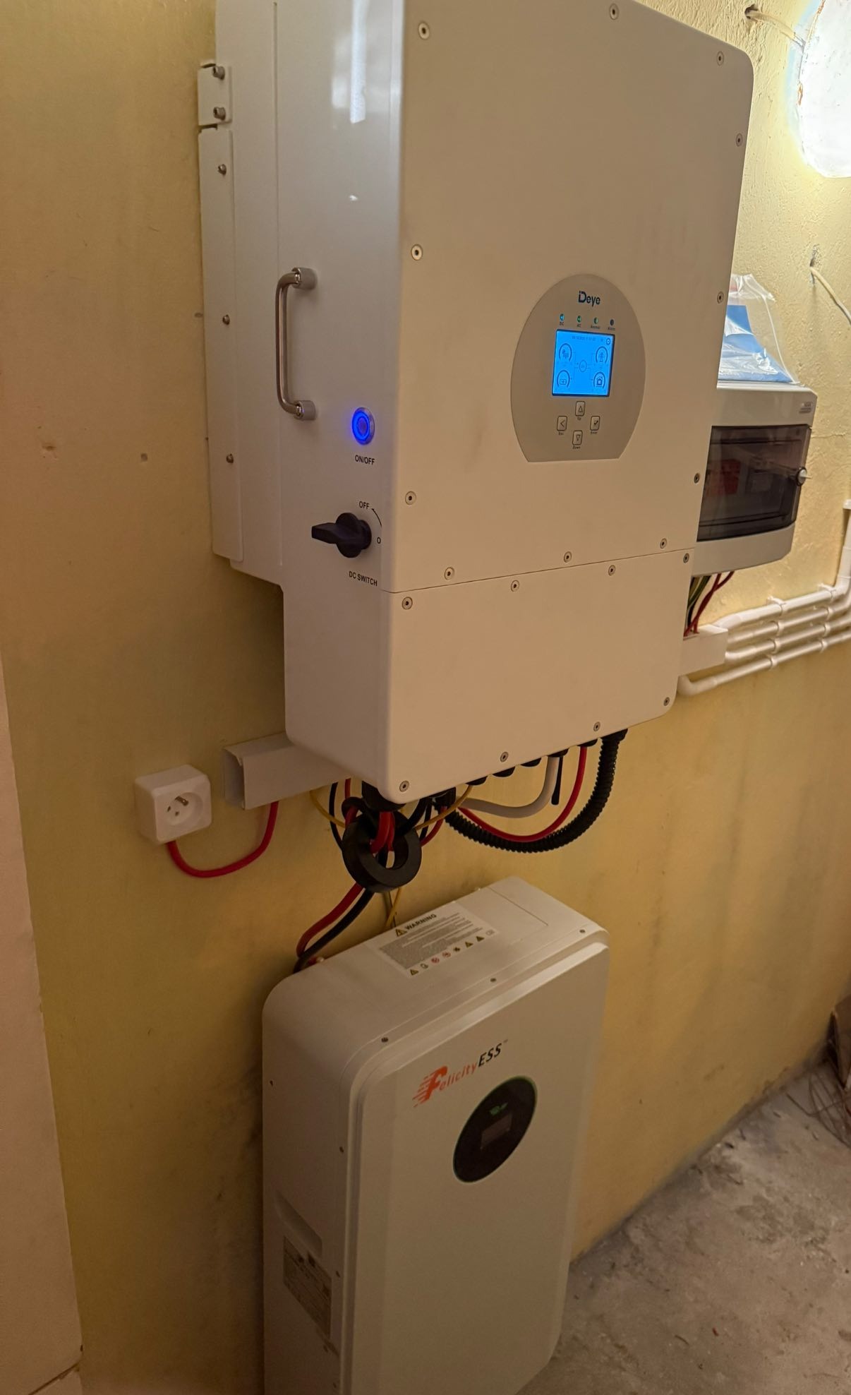 Zamontowany biały falownik Deye i bateria Felicity ESS na żółtej ścianie. Widoczne kable i elementy instalacji.