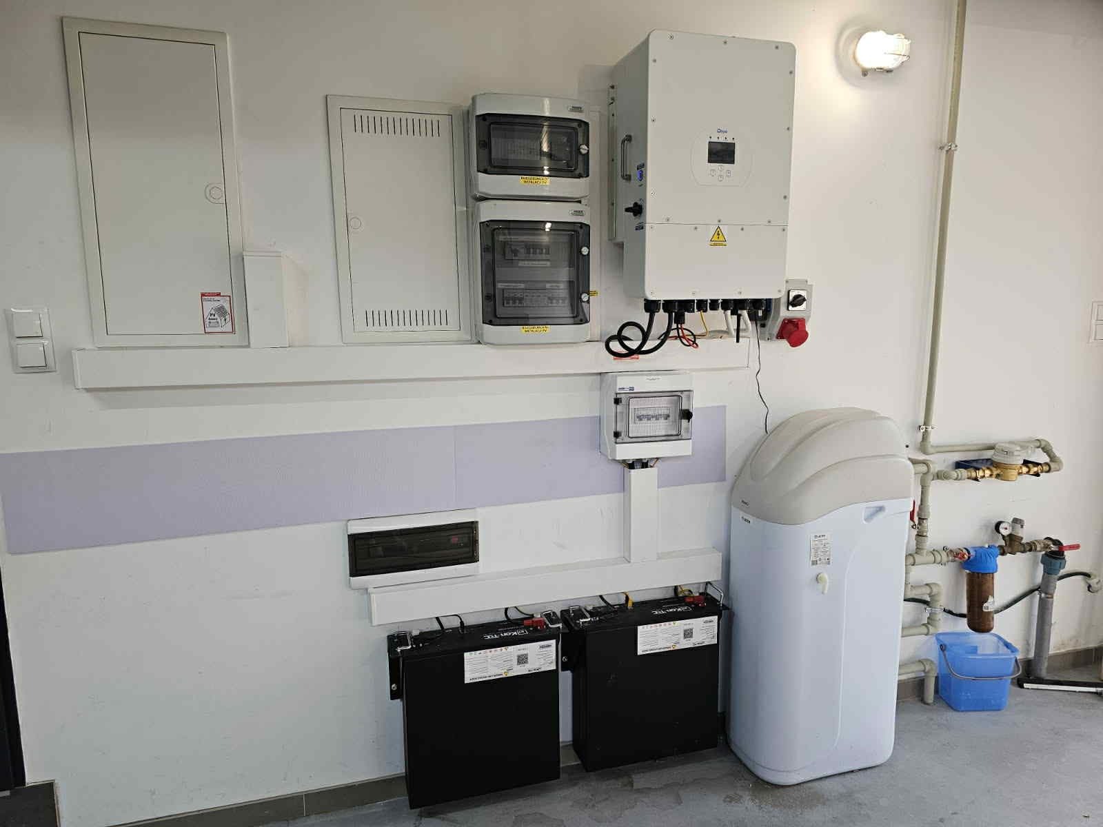Instalacja z inwerterem, rozdzielnicami, akumulatorami i zmiękczaczem wody na ścianie z fioletowym pasem. Widoczne przewody i elementy instalacji elektrycznej.
