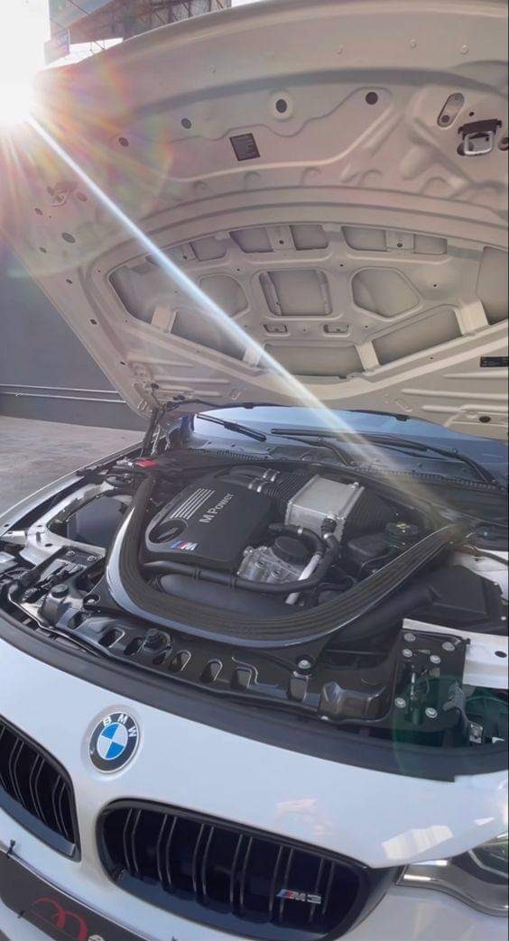 Otwarta maska białego samochodu BMW M3 z widocznym silnikiem M Power, oświetlona silnym światłem słonecznym.