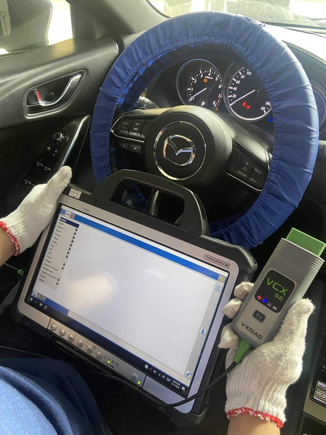 Diagnostyka samochodowa Mazda z użyciem laptopa Toughbook i interfejsu VXDIAG, widoczne rękawice ochronne i osłona na kierownicy.