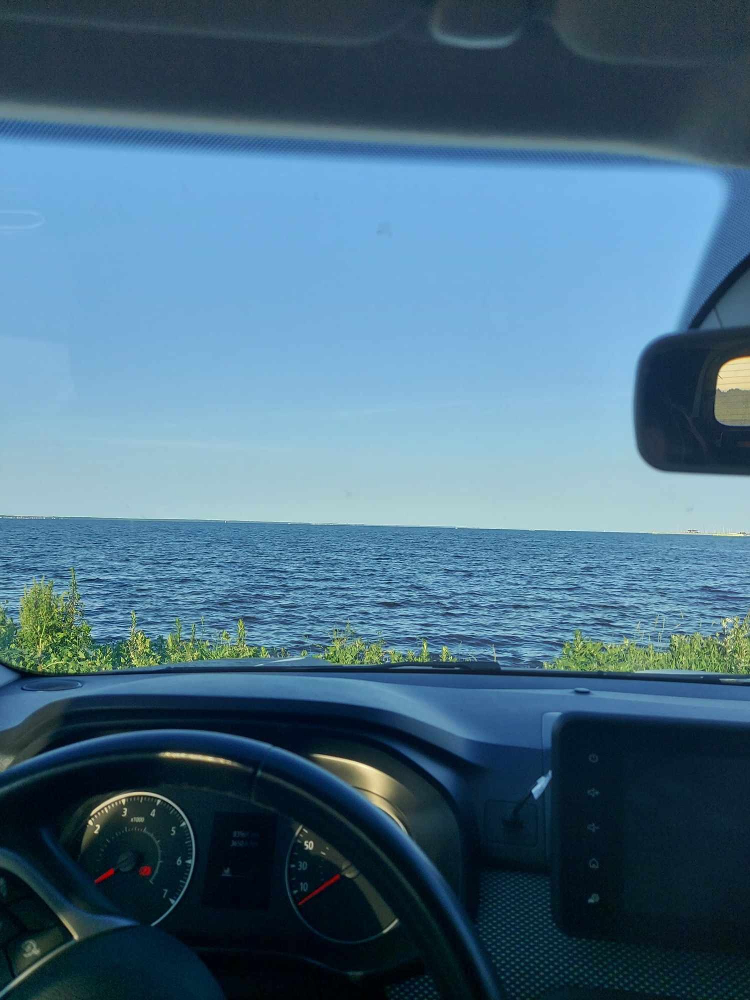 Widok z wnętrza samochodu na wodę i brzeg w słoneczny dzień, deska rozdzielcza z widocznymi zegarami i ekranem.