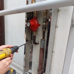 Radiator - Instalacja gazowa w osłonie ze stali nierdzewnej mocowana do ściany za pomocą wkrętarki DeWalt; widoczny reduktor ciśnienia, rury miedziane i poziomica na górnej krawędzi osłony.
