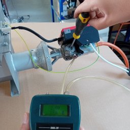 Radiator - Technik reguluje zawór gazowy śrubokrętem, jednocześnie monitorując ciśnienie gazu na elektronicznym manometrze EMS-20, w tle warsztatowy stół.