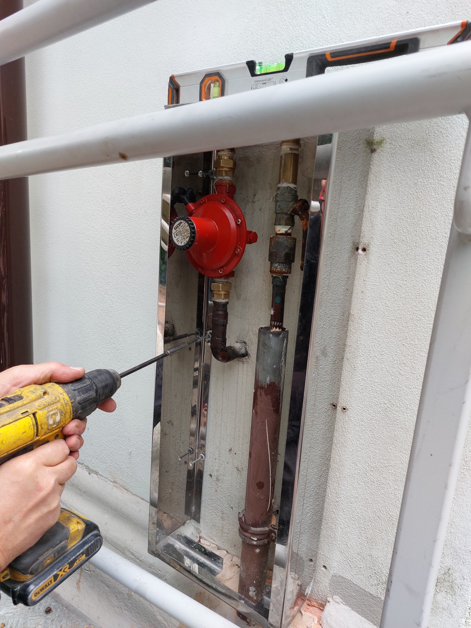 Instalacja gazowa w osłonie ze stali nierdzewnej mocowana do ściany za pomocą wkrętarki DeWalt; widoczny reduktor ciśnienia, rury miedziane i poziomica na górnej krawędzi osłony.