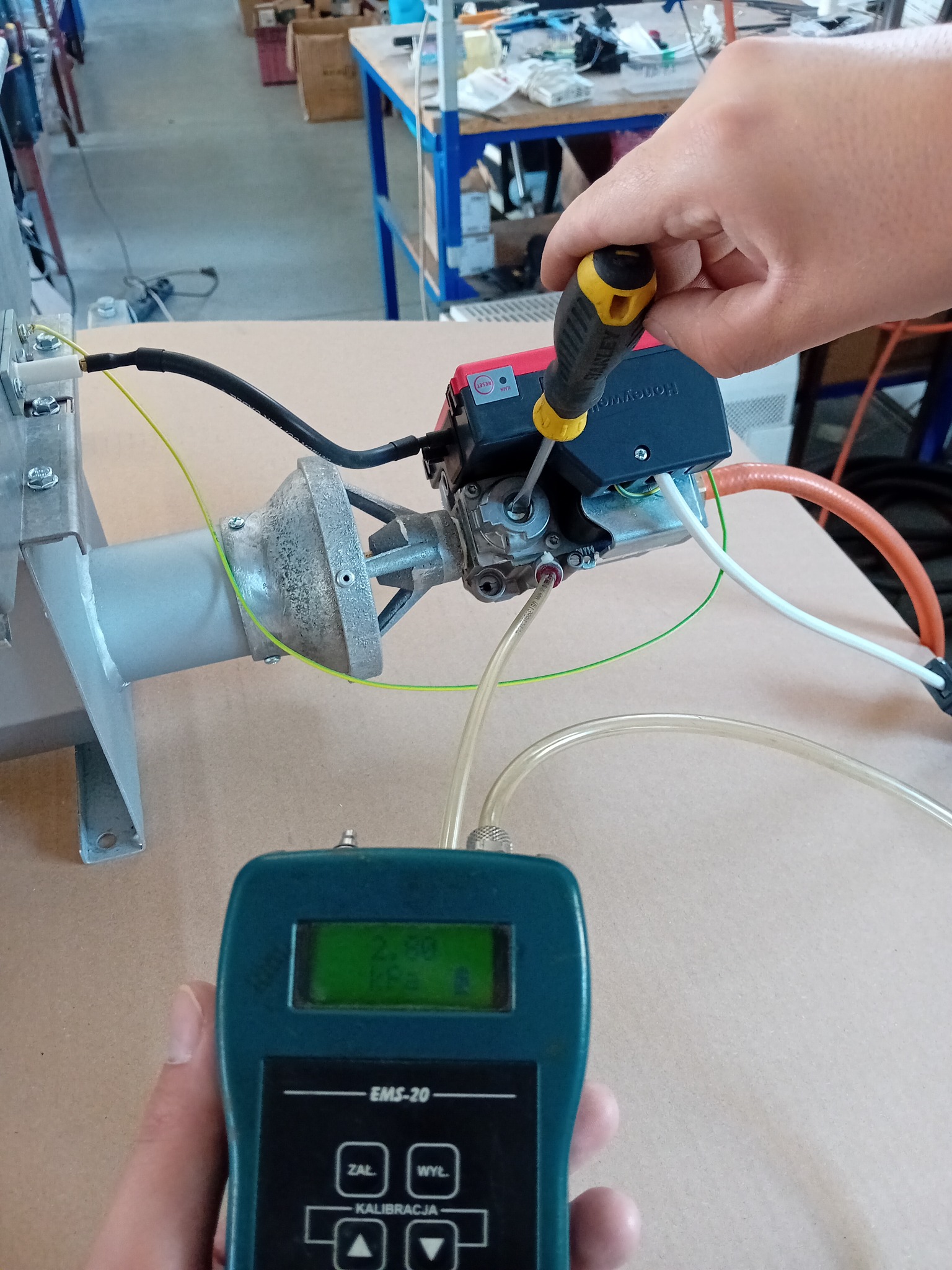 Technik reguluje zawór gazowy śrubokrętem, jednocześnie monitorując ciśnienie gazu na elektronicznym manometrze EMS-20, w tle warsztatowy stół.