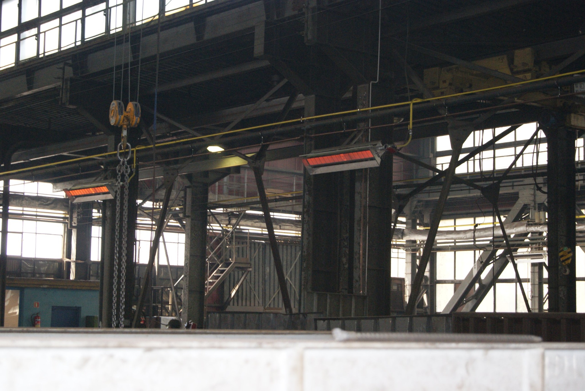 Wnętrze hali przemysłowej z widoczną suwnicą, stalowymi konstrukcjami, oknami i grzejnikami przemysłowymi.