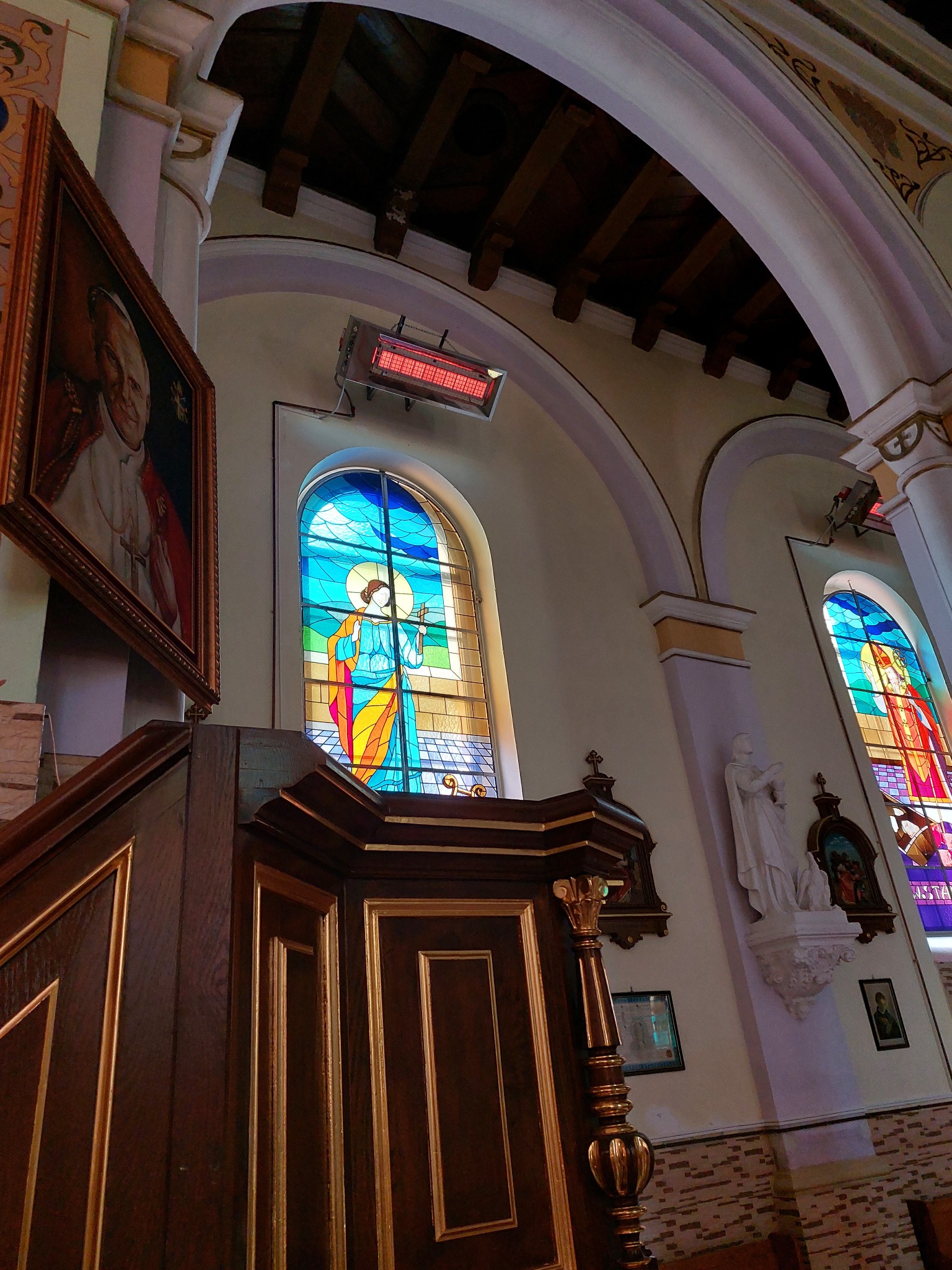Wnętrze kościoła z drewnianą amboną, witrażami w oknach i grzejnikiem elektrycznym pod sufitem.