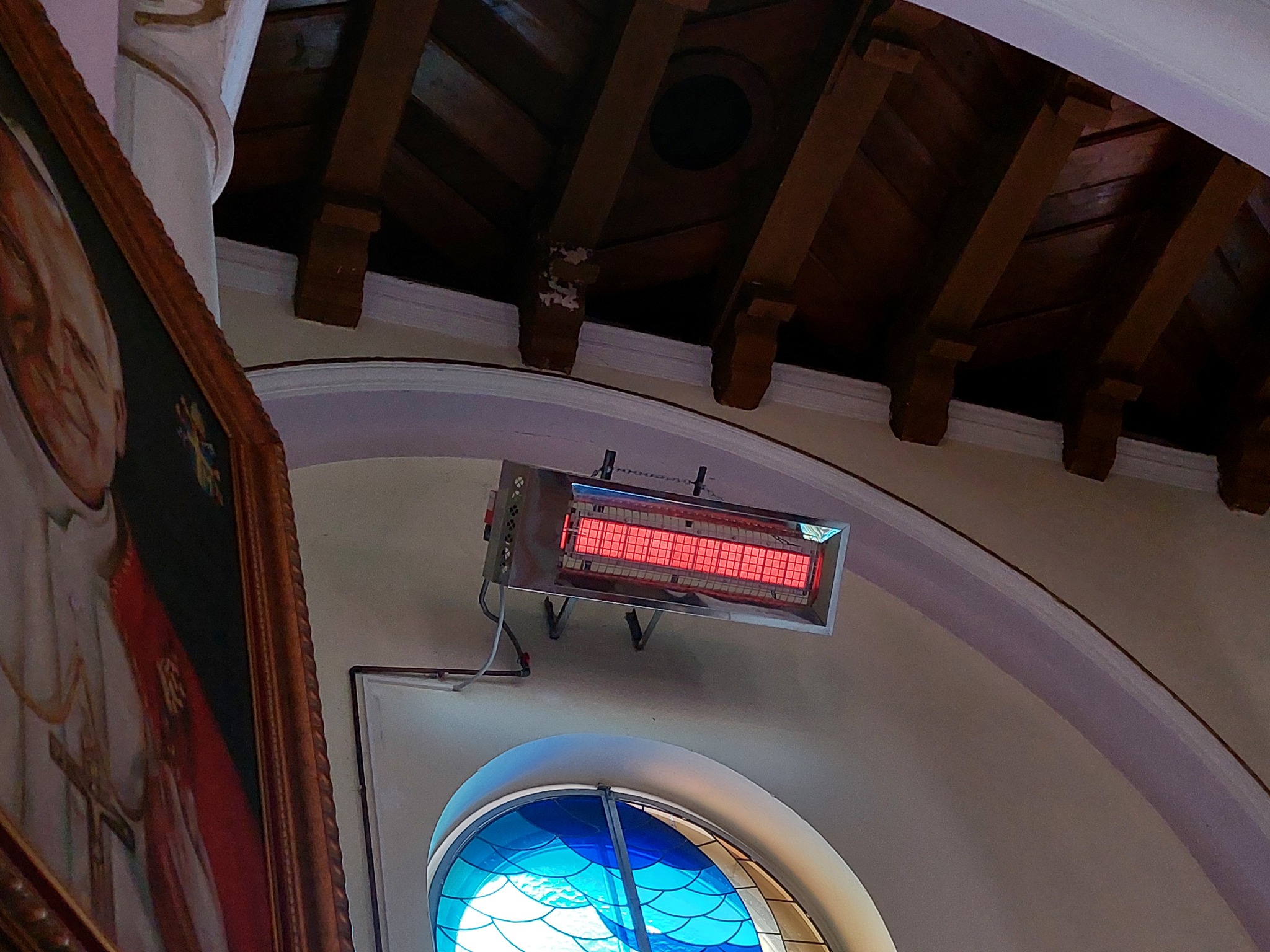 Ścienny grzejnik elektryczny z elementami grzejnymi w kolorze czerwonym, zamontowany wewnątrz budynku z drewnianym sufitem i witrażowym oknem w tle, obok obrazu w złotej ramie.