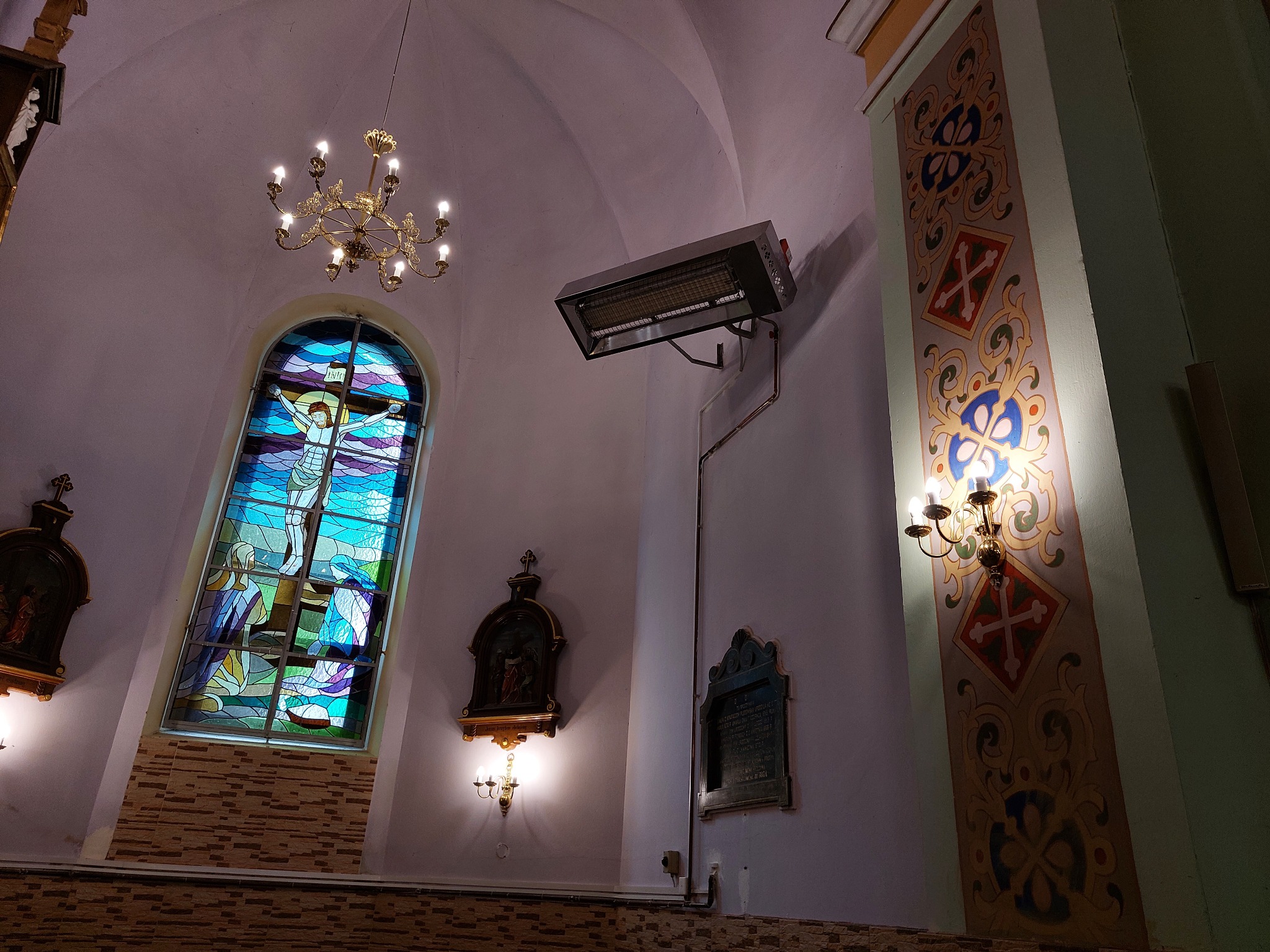 Wnętrze kościoła z witrażem, żyrandolem i grzejnikiem przemysłowym zamontowanym na ścianie za pomocą rur miedzianych.